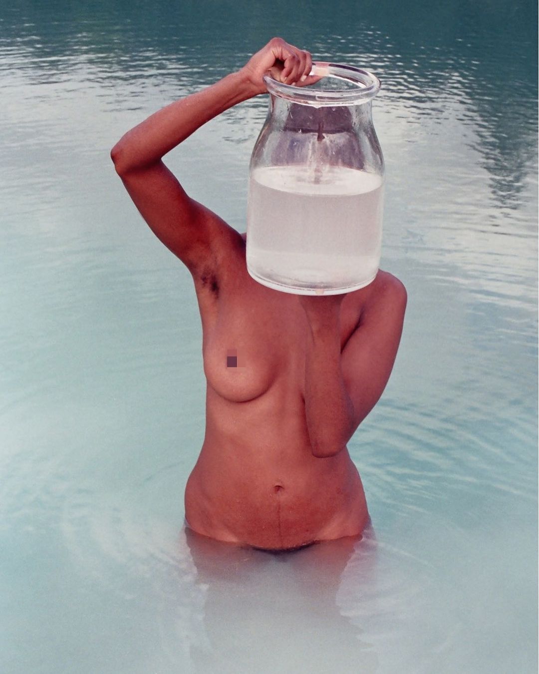 Nude adult stands in blue water holding large glass jar over head, obscuring the face in this conceptual outdoor art photo.