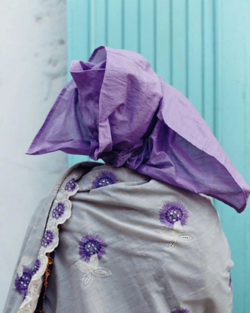 Woman wearing a traditional gray embroidered shawl and light purple head covering, standing against a turquoise and white wall.
