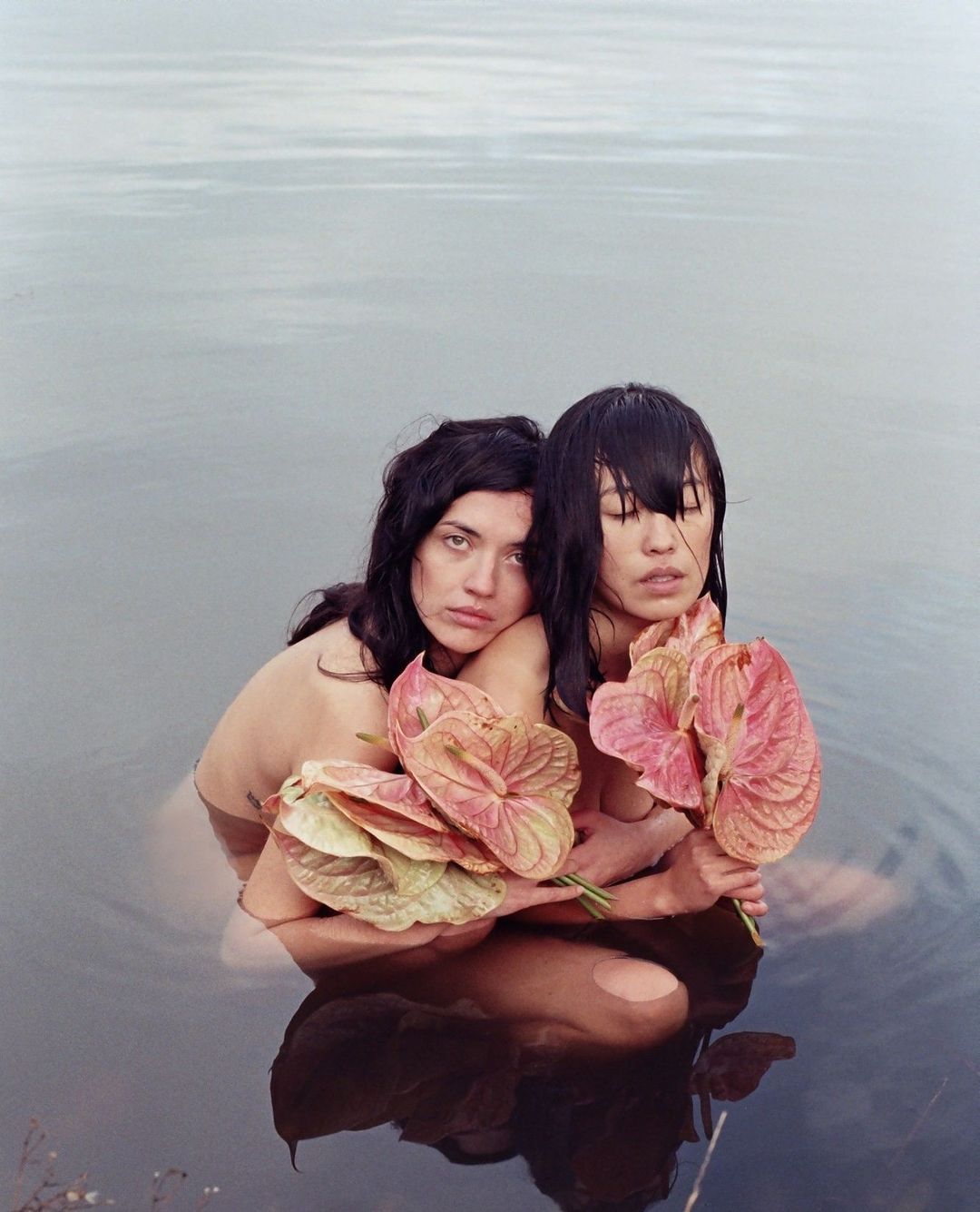 Two women stand in water holding anthurium flowers with pink and green spathes, creating a serene and artistic composition with a tranquil backdrop.