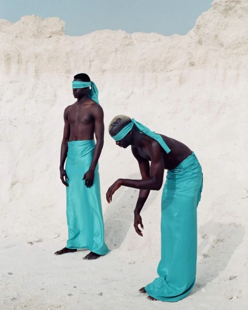 Two dark-skinned adults are outdoors wearing turquoise fabric wraps as blindfolds and skirts, standing in a bright, sandy environment.
