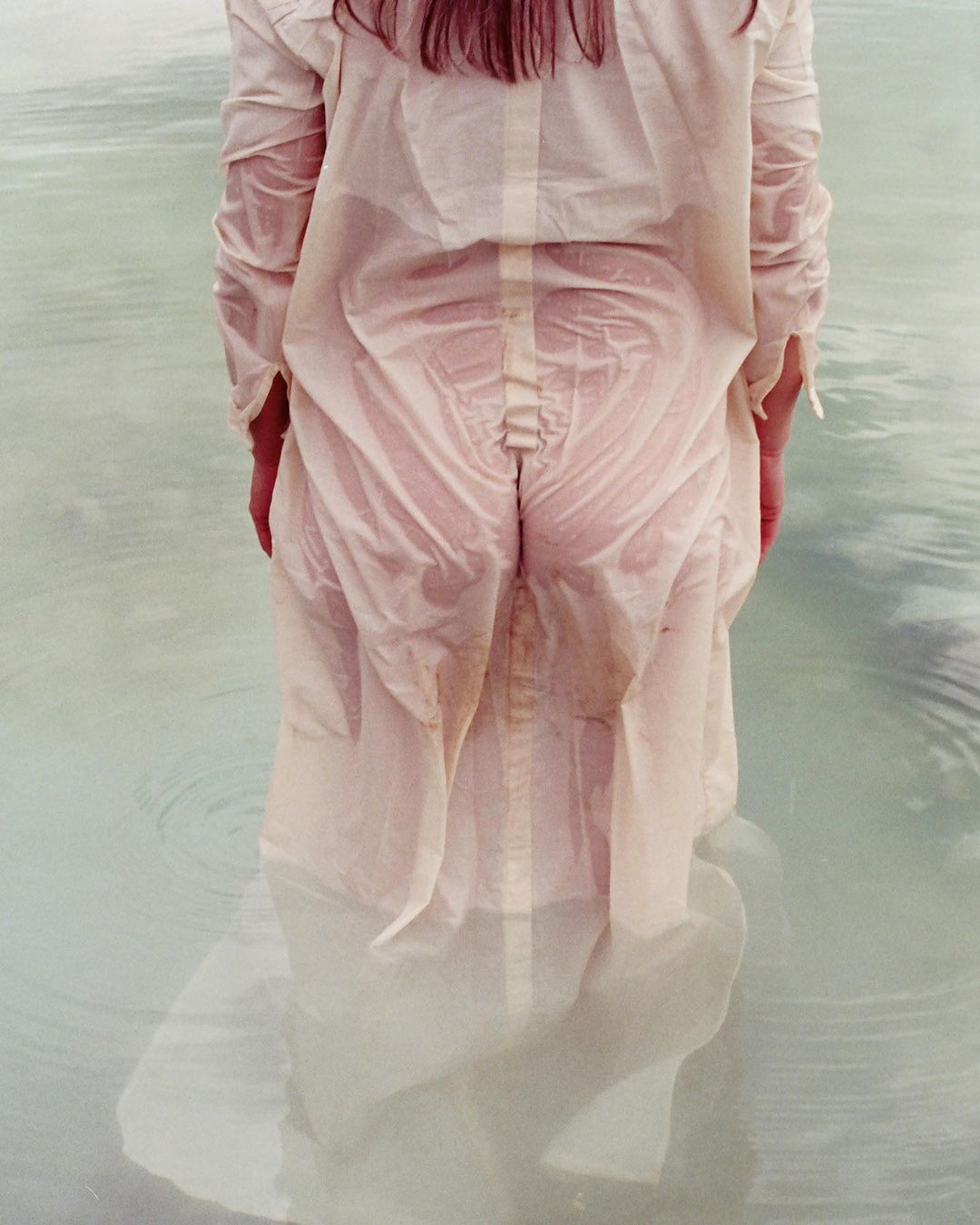 A woman in a wet white dress stands in shallow water, creating ripples and reflections, in a minimalist, artistic fashion photograph.
