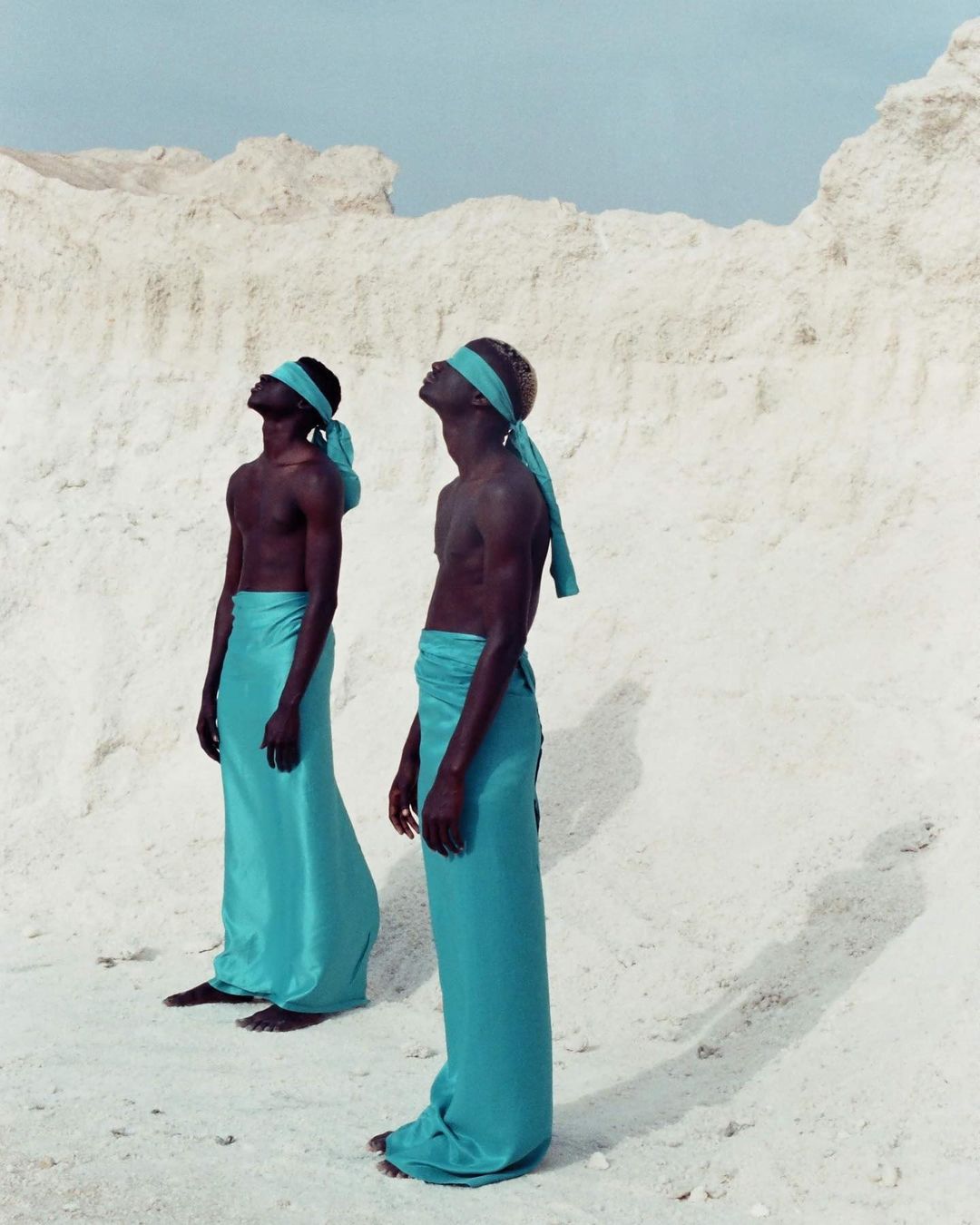 Two men in matching turquoise wraps stand on white sand with heads tilted back in an outdoor, dreamlike fashion shot.