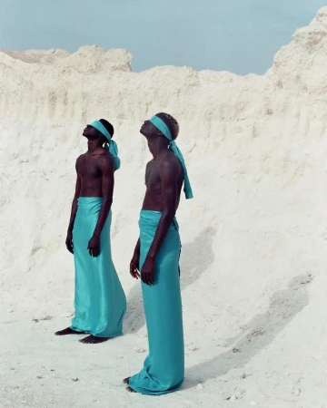 Two men in matching turquoise wraps stand on white sand with heads tilted back in an outdoor, dreamlike fashion shot.