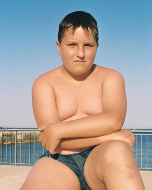 Child posing shirtless at a sunny beach with blue sky. Boy seated outdoors in swimwear, arms crossed. Seascape background with clear skies.