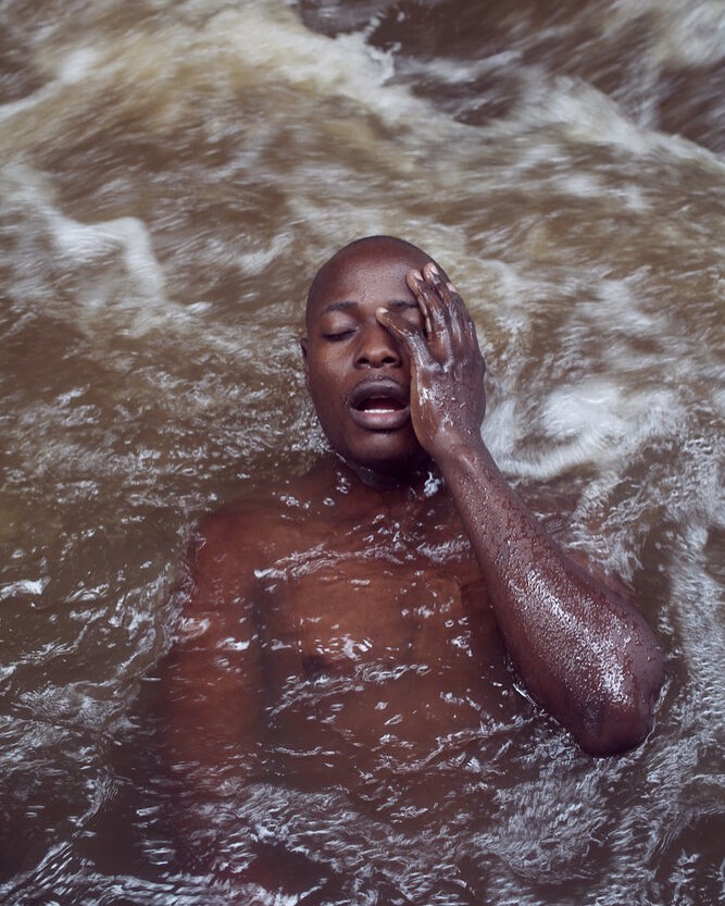 A man with dark skin is submerged in rushing, foamy water with his hand on his face. The water is brown and the man's eyes are closed.