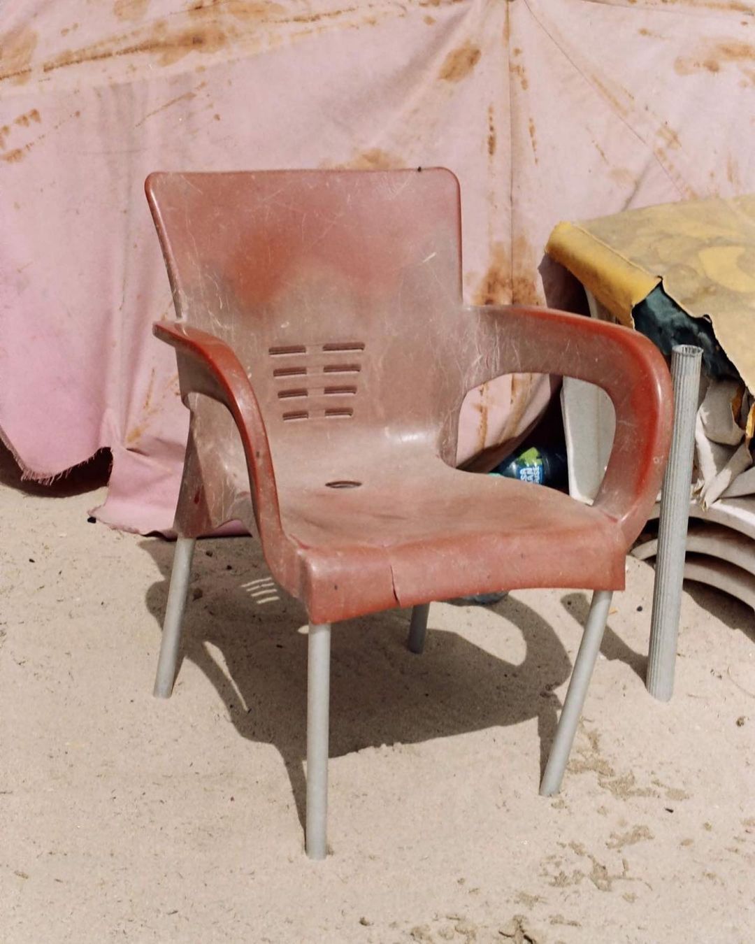 A red, worn plastic chair with aluminum legs sits on a sandy surface, with a pink and white patterned tarp in the background.