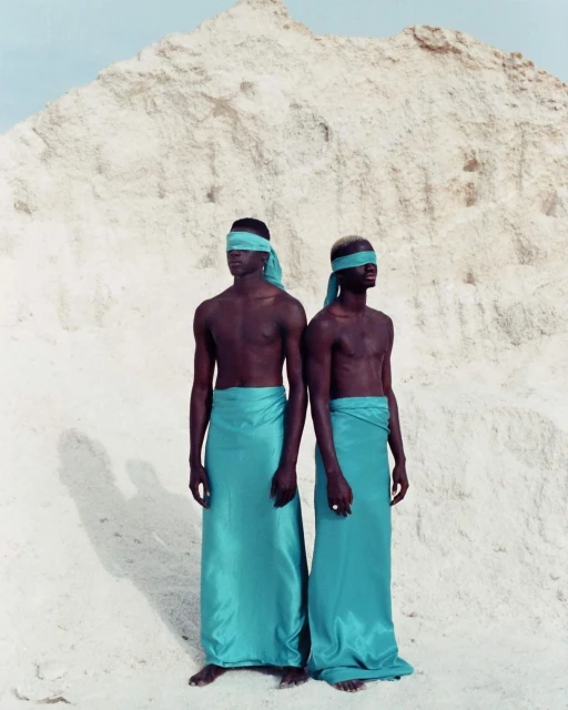 Two dark-skinned adult men stand blindfolded, wearing aqua-colored sarongs in a white sandy landscape with a large sand dune behind.