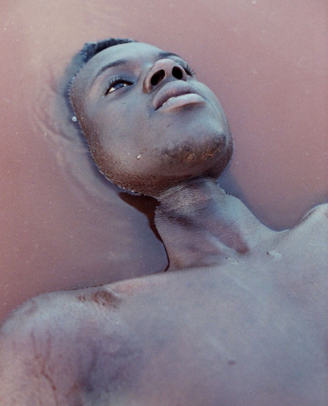 Close-up portrait of a Black adult male partially submerged in calm, reddish-brown water, face tilted upward looking off-frame, outdoors.