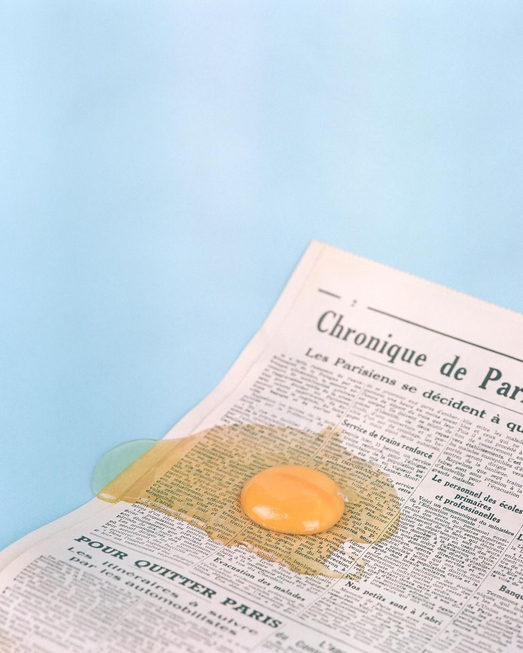 Close-up of a cracked raw egg with yellow yolk, sitting on a vintage French newspaper against a light blue background, studio lighting.