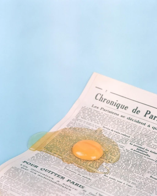 Close-up of a cracked raw egg with yellow yolk, sitting on a vintage French newspaper against a light blue background, studio lighting.