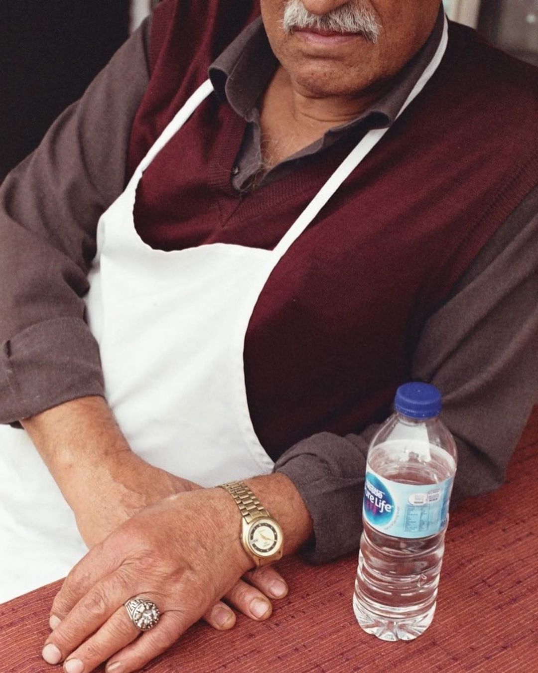 Man wearing apron and gold watch rests hands near water bottle on red table. Photo of restaurant worker in candid indoor setting.