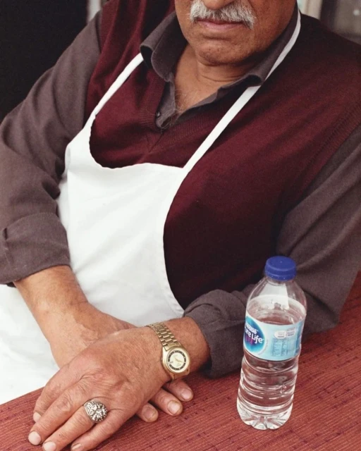 Man wearing apron and gold watch rests hands near water bottle on red table. Photo of restaurant worker in candid indoor setting.