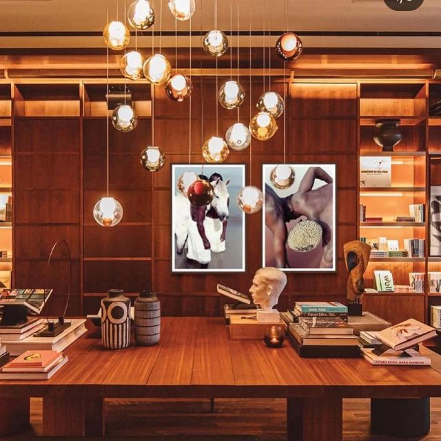 Stylish interior featuring a wooden table with books, vases, and sculptures, framed by artwork and decorative lighting with warm tones and bookshelves.
