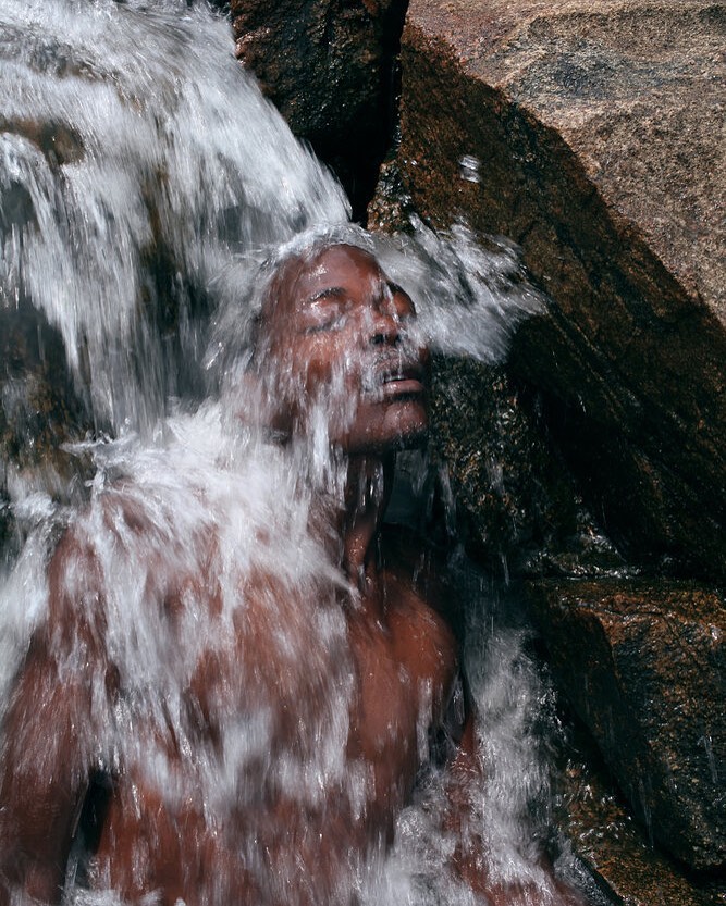 African American man under a waterfall with water cascading over his face and body, surrounded by dark, wet rocks in a natural setting.