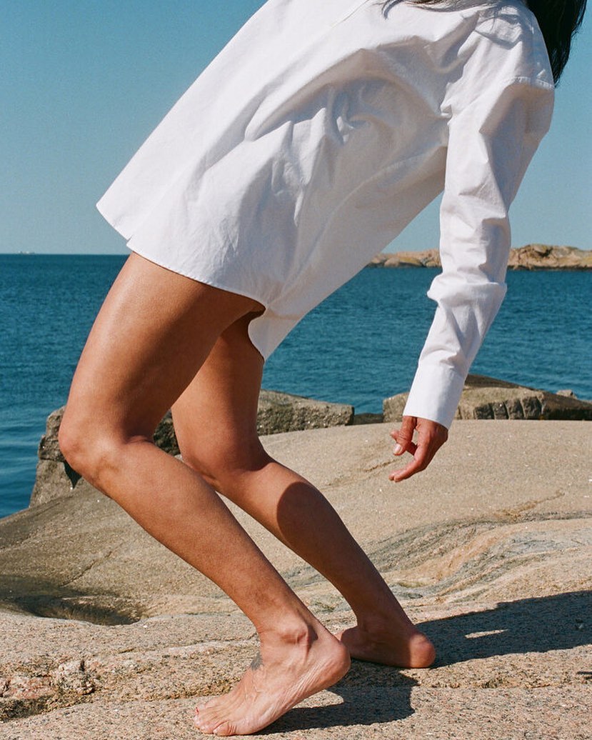 Barefoot adult woman stands on a rock near the ocean wearing a white shirt. The sky is clear and blue in an outdoor setting.