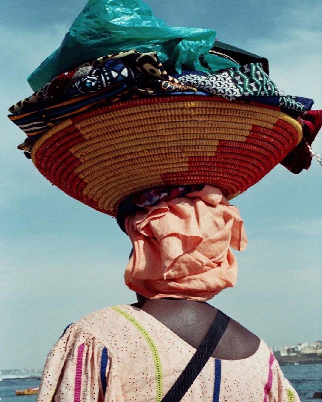 Woman carrying a large woven basket filled with fabrics on her head near a coastal area under a bright sky.
