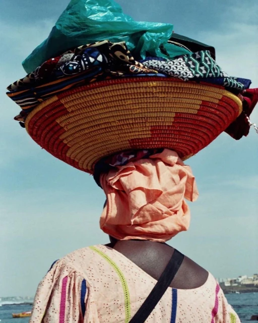 Woman carrying a large woven basket filled with fabrics on her head near a coastal area under a bright sky.