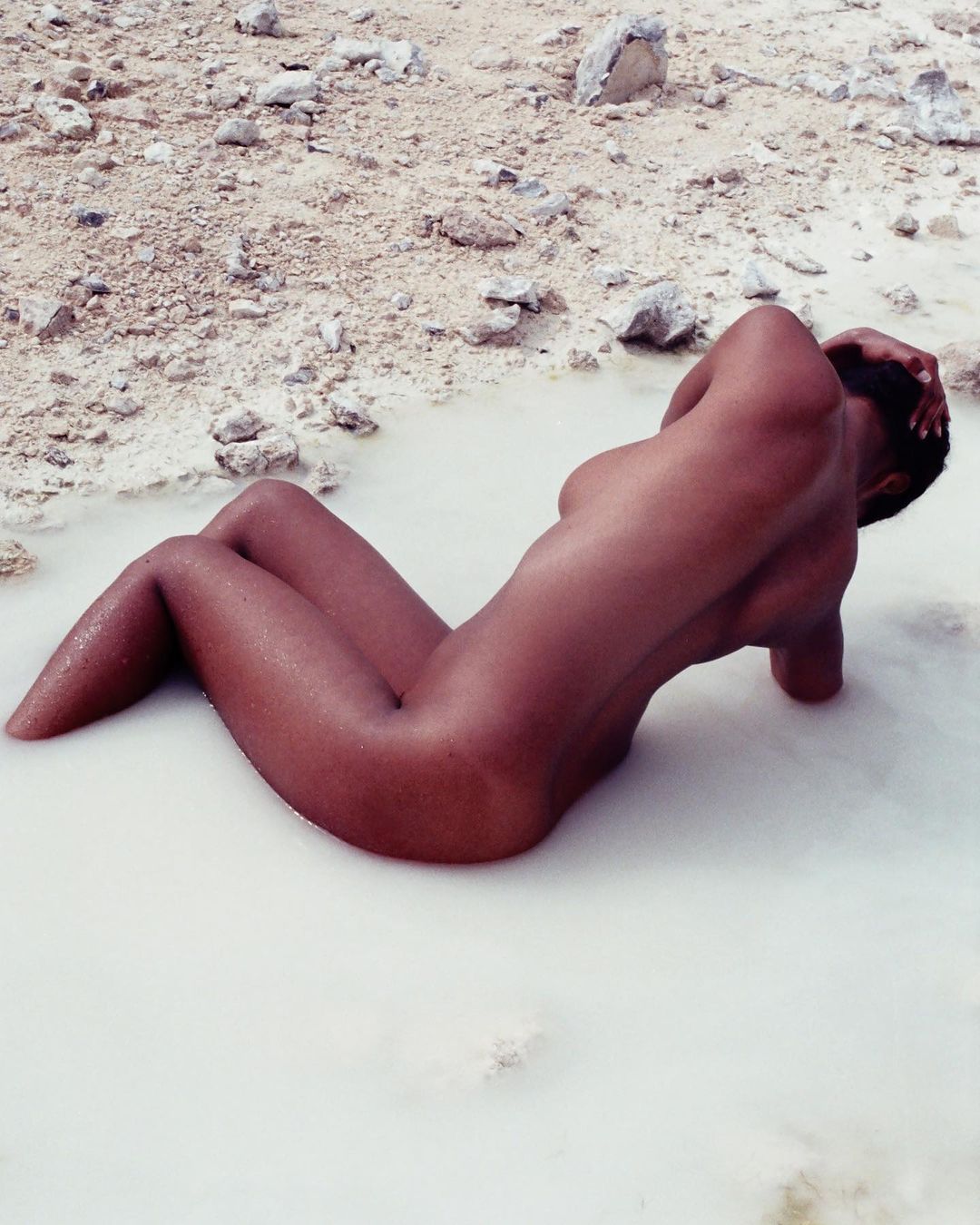 Nude adult female lying in a body of water surrounded by light coloured rocks, angled portrait orientation, neutral facial expression.