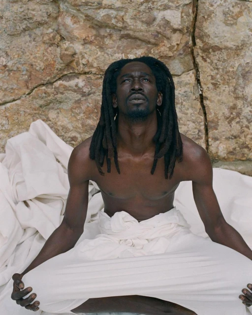 Man with dreadlocks, shirtless, wearing white pants, sitting cross-legged against a rocky backdrop in an outdoor setting, looking upward.