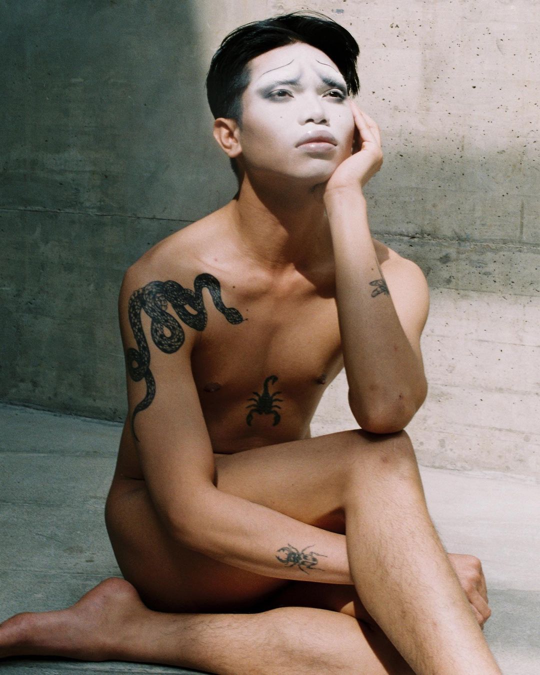 Nude figure with face makeup sitting against concrete wall, showcasing snake, scorpion, beetle tattoos, neutral tones, studio lighting.