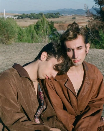 Two men in brown leather jackets outdoors with one resting his head against the other in a rural landscape by Denisse Ariana Pérez