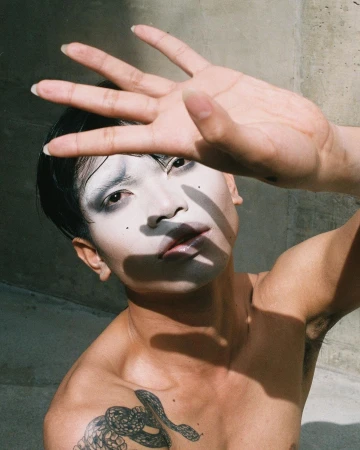 Person with dramatic white makeup and a snake tattoo shields their face from the sun near a concrete wall outdoors.