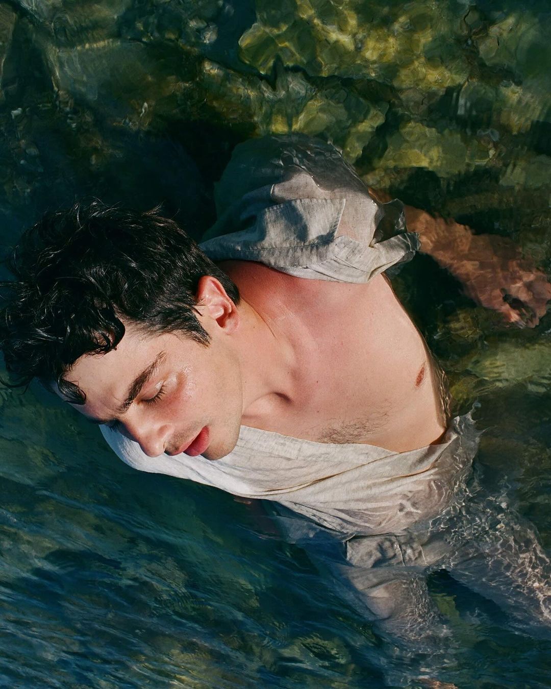 A shirtless man in a gray linen shirt partially submerged in clear water, eyes closed, rocks visible through the water.