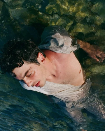 A shirtless man in a gray linen shirt partially submerged in clear water, eyes closed, rocks visible through the water.