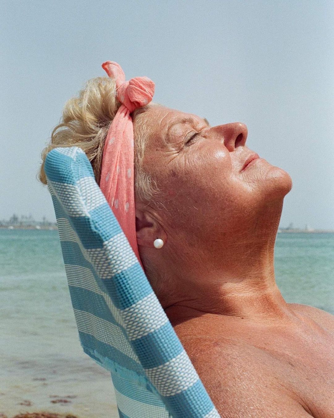Mature woman relaxes in a striped deckchair by the ocean, wearing a pink polka-dot headscarf and white earrings on a sunny day.