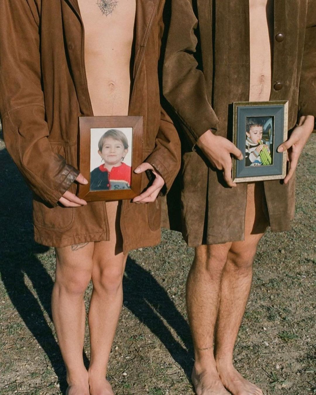 Two barefoot adults wearing brown coats hold framed child photos in an outdoor setting with sparse ground cover and natural light.