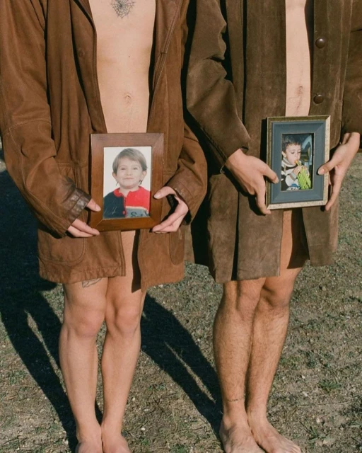 Two barefoot adults wearing brown coats hold framed child photos in an outdoor setting with sparse ground cover and natural light.