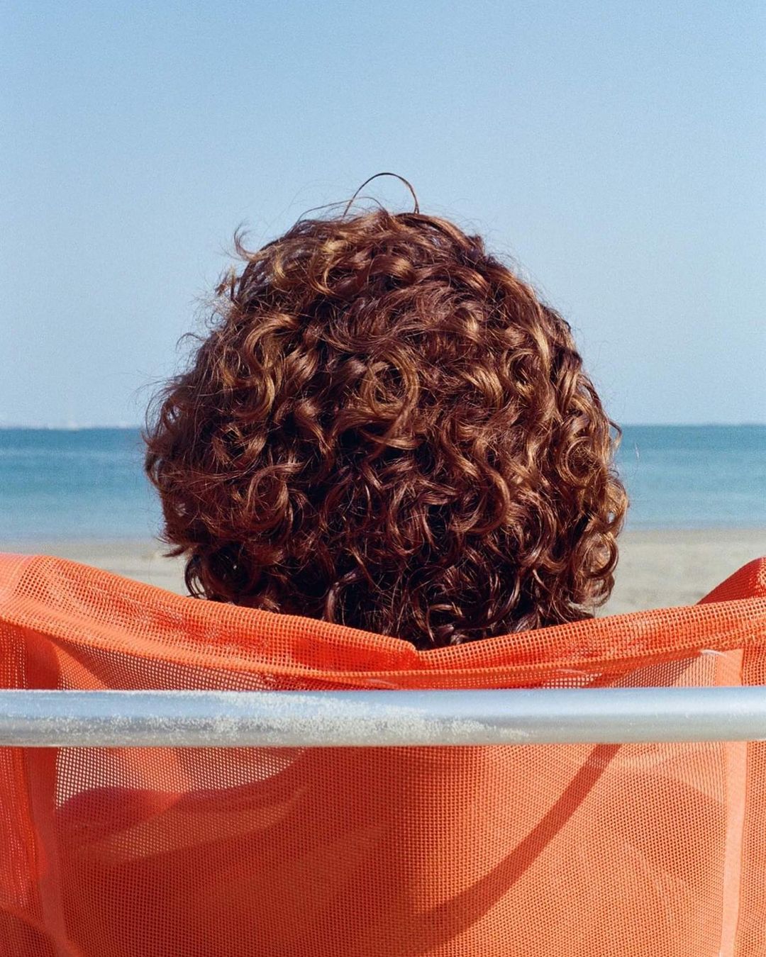 Person with curly hair sits in orange chair at the beach. Clear blue sky and water are visible in the background.