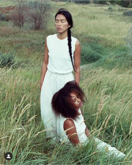 Two women in sleeveless tops and light pants pose in a grassy field, one standing and one seated, with soft lighting and a natural setting.
