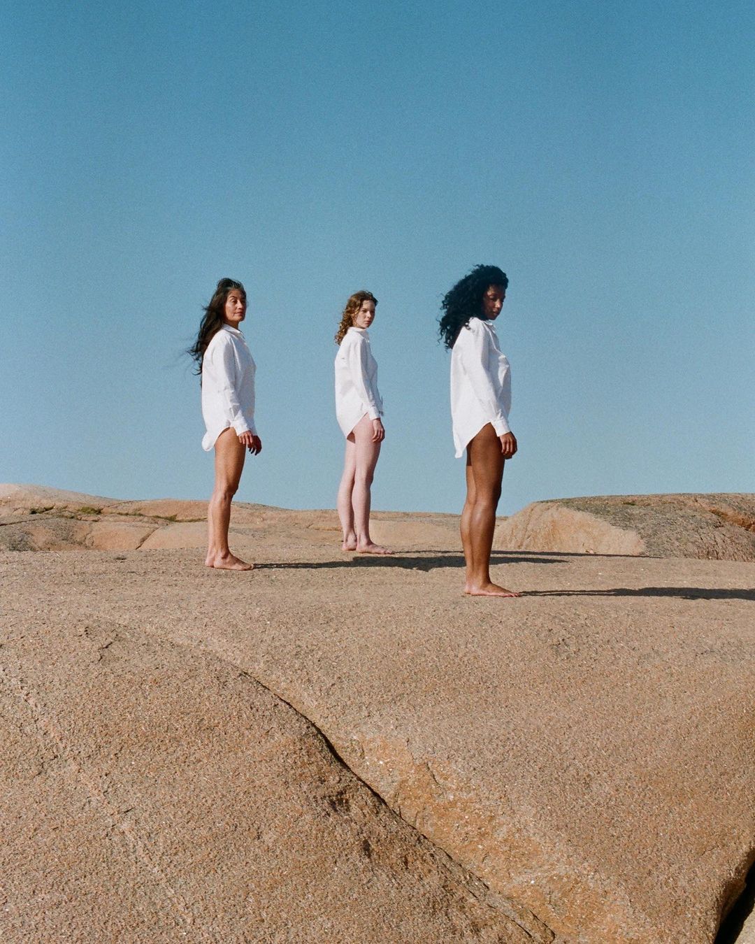 Three women stand in white shirts on a rock formation against a clear blue sky, creating a minimalist and serene outdoor scene.