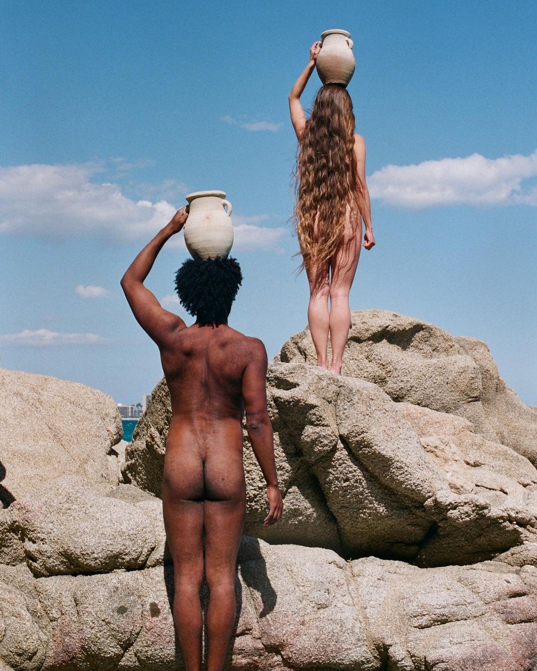 Two nude figures on rocks, each balancing a clay pot on their head against a partly cloudy sky.