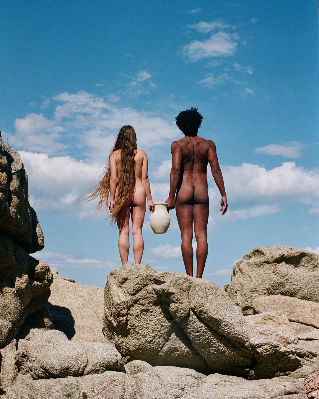 Two nude figures, an adult female and adult male, stand on rocks holding a jug against a blue sky with light clouds.