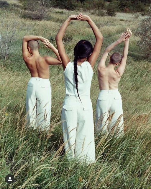 Three figures in white pants stand in a grassy field with arms raised, forming circles with their arms and hands under natural light.