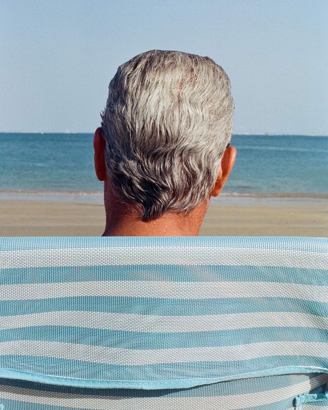 Elderly man with gray hair sits on a blue and white striped beach chair looking out at the ocean and sandy beach on a bright sunny day.