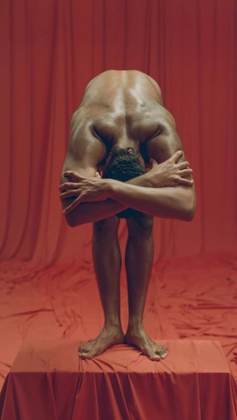 Muscular adult male with head bent down, arms crossed, standing on a red table against a matching red background in a studio.