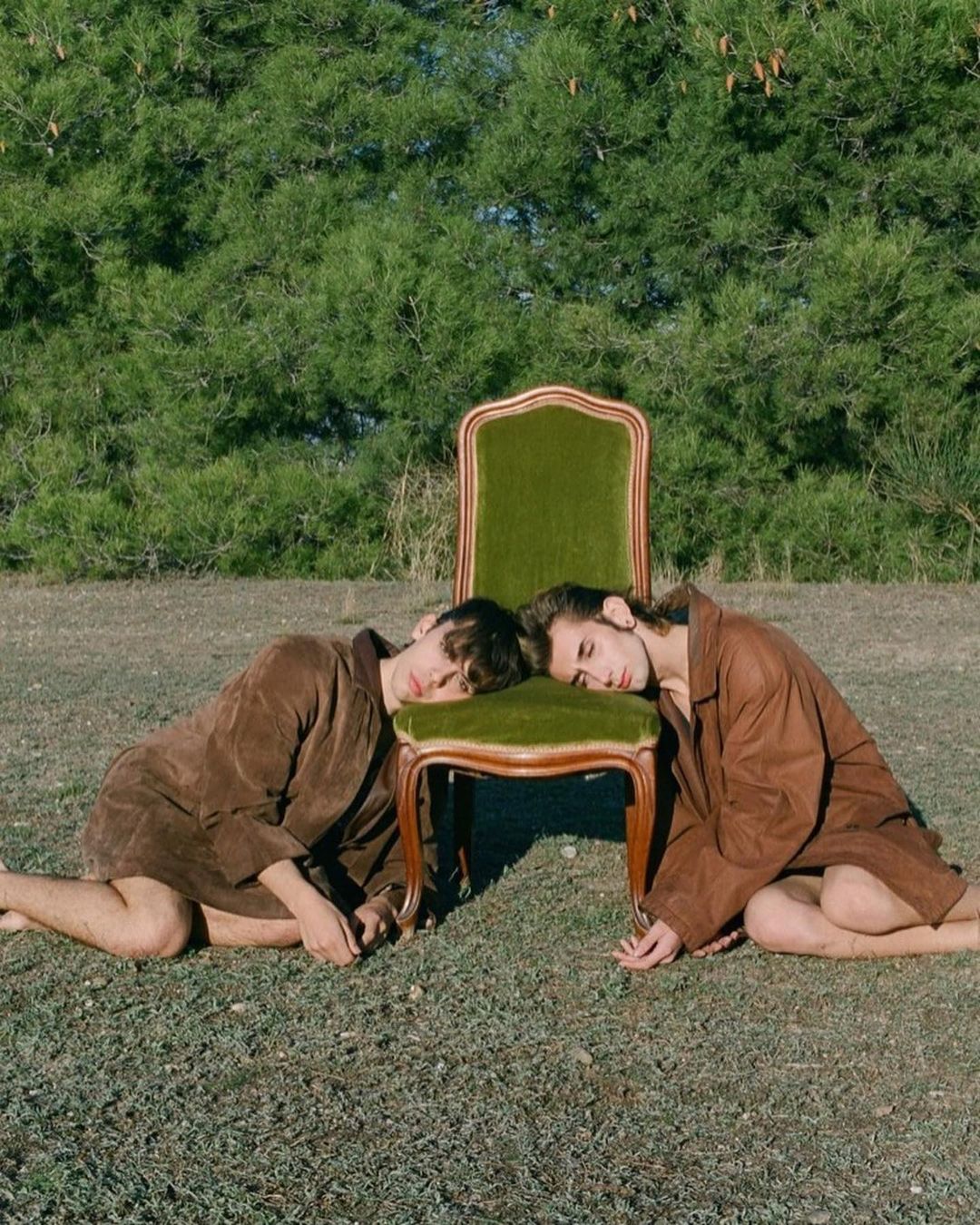 Two people in brown robes resting their heads on a green vintage chair in a grassy field with trees in the background on a sunny day.