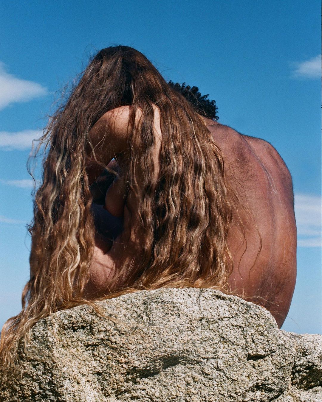 Back view of two people with long hair sitting together on a rock under a bright blue sky. Brown and tan skin tones visible.