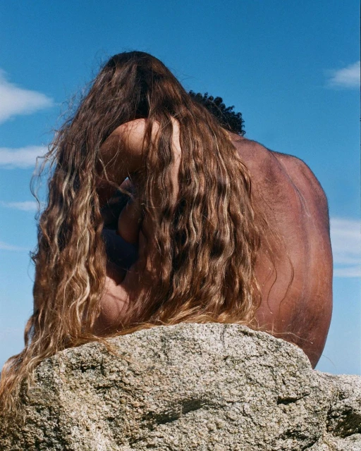 Back view of two people with long hair sitting together on a rock under a bright blue sky. Brown and tan skin tones visible.