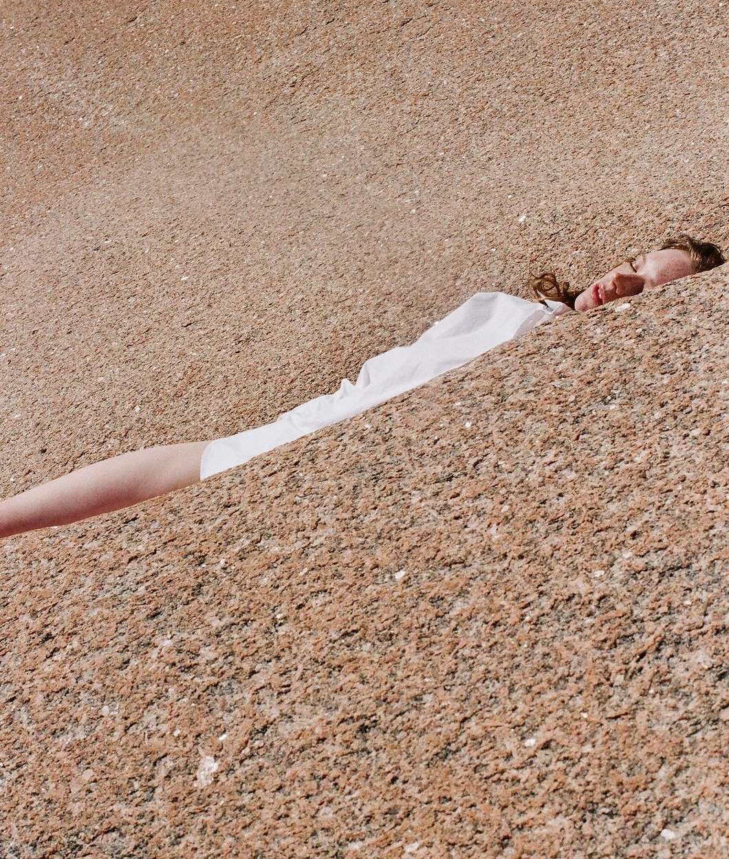 Adult woman wearing a white shirt lies on a granular, tan-colored surface in direct sunlight; only the face, neck, and part of the body are visible.