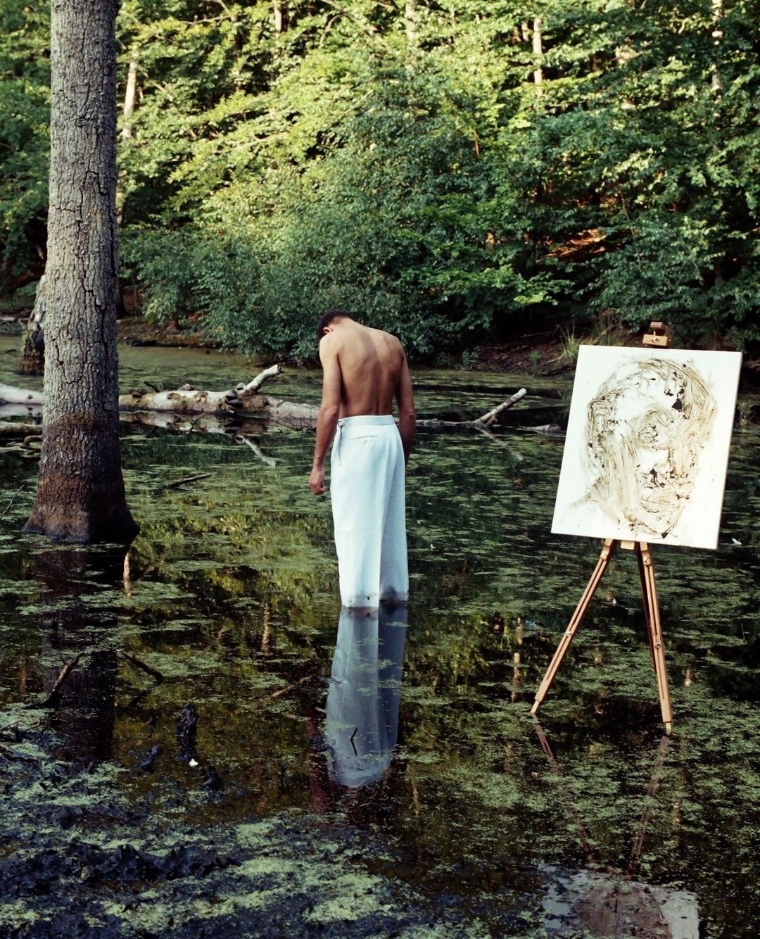 Man standing in swamp in front of easel with abstract painting, surrounded by trees and foliage with reflections in the water.