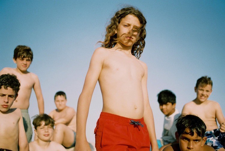 A group of young boys pose outdoors against a clear sky, with one in red swim trunks dominating the foreground in a medium shot.