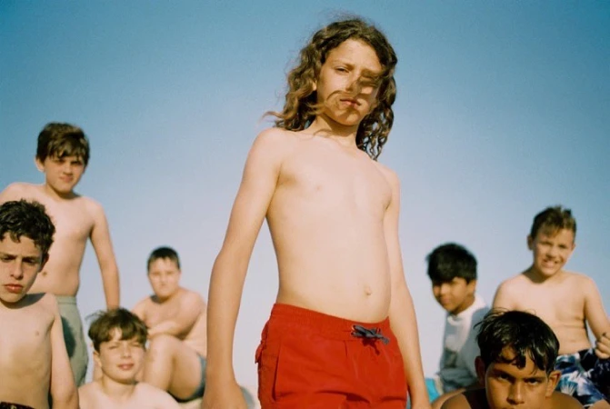 A group of young boys pose outdoors against a clear sky, with one in red swim trunks dominating the foreground in a medium shot.