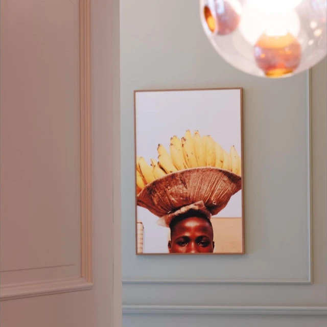 Photo of an art print of a person balancing a basket of bananas on their head, mounted on a pale blue wall in a neutral room.
