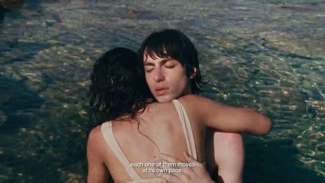 Two people embracing in shallow coastal water with subtitle text overlay at the bottom