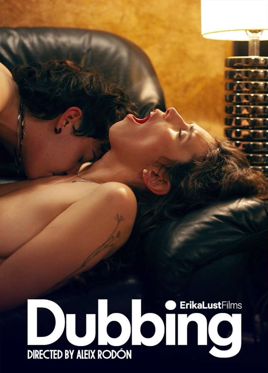 Two adult women are intimately close on a black leather couch, one with their mouth open. A decorative lamp is in the background.