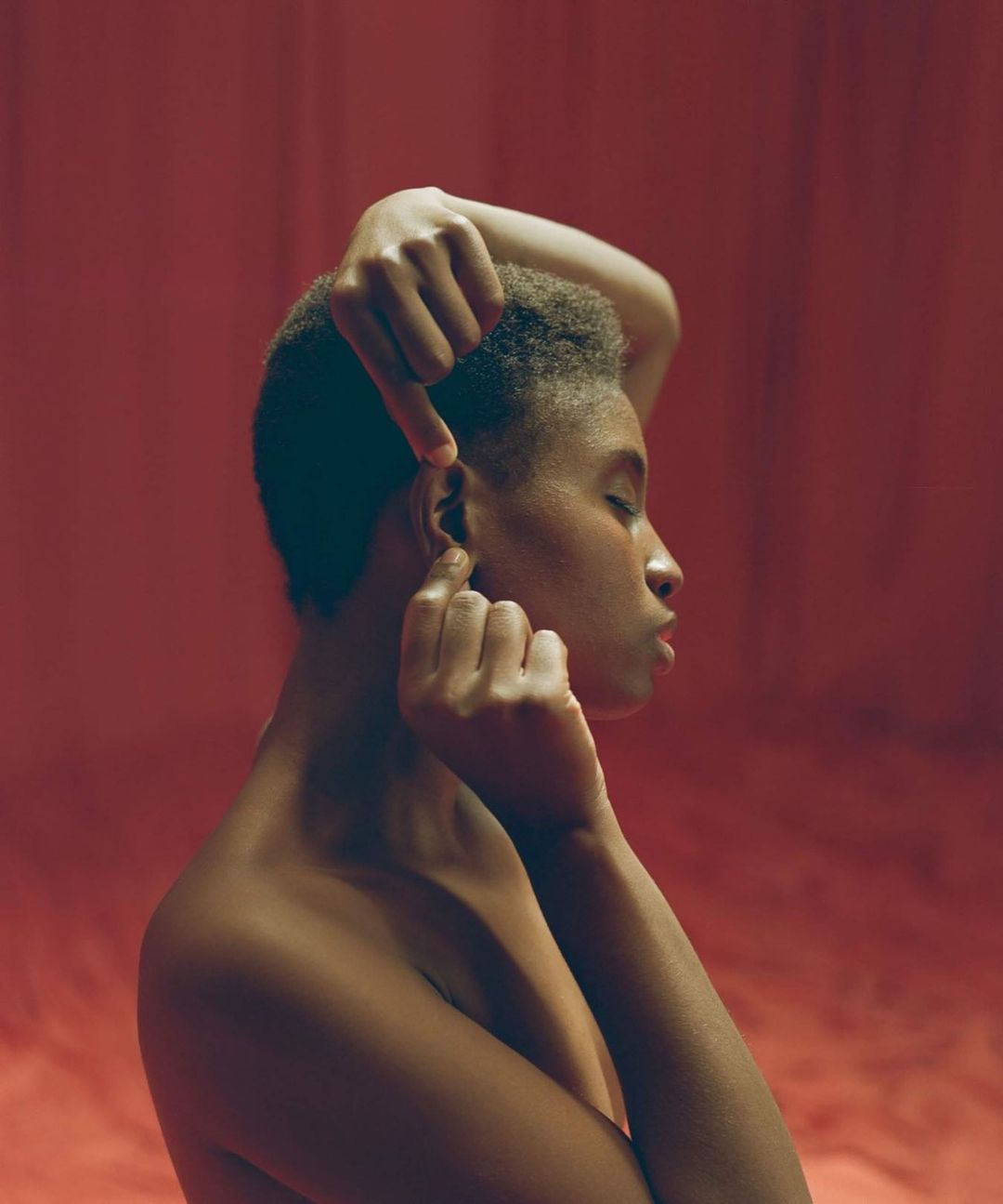 Portrait of a Black woman with short hair holding her ear against a rich red background by Denisse Ariana Pérez